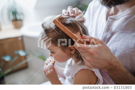 Unrecognizable father with small daughter indoors in bathroom at home, combing hair. 83131803