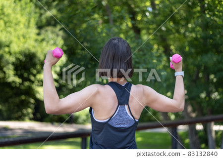 Fitness trainer conducts workout in the park. Girl holds small dumbbells and does the exercise. Back view. Fitness trainer conducts workout in the park. Girl holds small dumbbells and does the exercise. Back view. 83132840