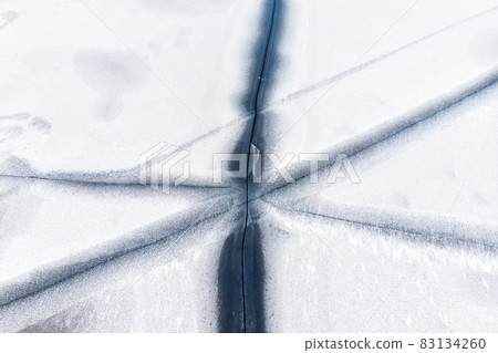 Aerial drone top view of snow covered frozen lake or river surface with big cracked ice diagonal lines. Natural winter landscape abstract texture pattern. Dangerous pond melting at thaw season 83134260