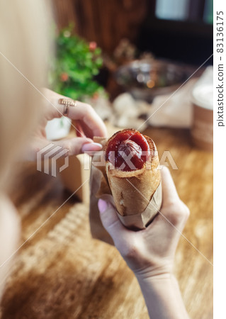 Close-up of a hot dog in hand. The girl has a French manicure. There is a lot of ketchup on top and sauce on the food. The woman wrapped her hands around the bun, creating a hint of intimacy. 83136175