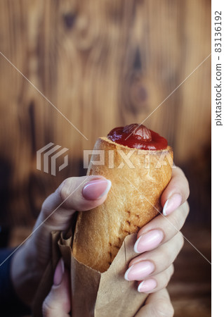 Close-up of a hot dog in hand. The girl has a French manicure. There is a lot of ketchup on top and sauce on the food. The woman wrapped her hands around the bun, creating a hint of intimacy. Close-up of a hot dog in hand. The girl has a French manicure. There is a lot of ketchup on top and sauce on the food. The woman wrapped her hands around the bun, creating a hint of intimacy. 83136192