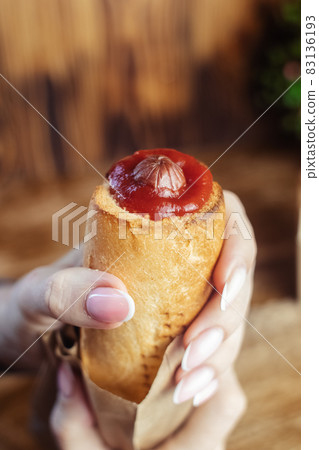 Close-up of a hot dog in hand. The girl has a French manicure. There is a lot of ketchup on top and sauce on the food. The woman wrapped her hands around the bun, creating a hint of intimacy. Close-up of a hot dog in hand. The girl has a French manicure. There is a lot of ketchup on top and sauce on the food. The woman wrapped her hands around the bun, creating a hint of intimacy. 83136193