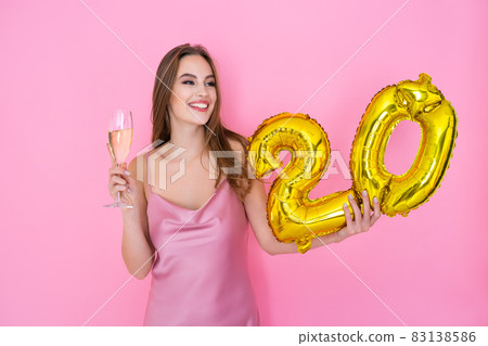 Young happy girl holds gold foil balloon and glass of champagne on pink background. BIRTHDAY PARTY 83138586