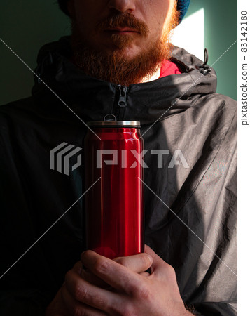 Red thermo can bottle in male hands against wall with harsh shadow.Hot tea coffee drinking in cold autumn winter weather Red thermo can bottle in male hands against wall with harsh shadow.Hot tea coffee drinking in cold autumn winter weather 83142180