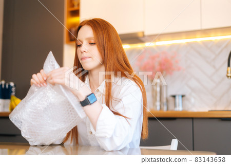 Close-up face of stressed young woman playing pops bubble wrap to calm herself sitting at table in kitchen. Close-up face of stressed young woman playing pops bubble wrap to calm herself sitting at table in kitchen. 83143658
