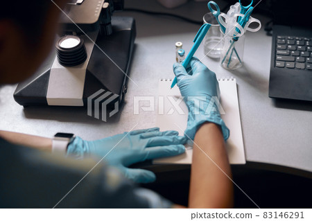 Woman in gloves writes in notebook near microscope at table in laboratory Woman in gloves writes in notebook near microscope at table in laboratory 83146291
