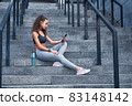 Young athletic woman in sport clothes sitting on stairs of city stadium with phone in hands posting her training results 83148142