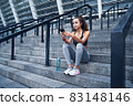 Young athletic woman in sport clothes sitting on stairs of city stadium with phone in hands posting her training results 83148146