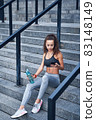 Young athletic woman in sport clothes sitting on stairs of city stadium with phone in hands posting her training results 83148149