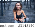 Young athletic woman in sport clothes smiling and listening to music while sitting on stairs of city stadium 83148150