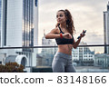 Young athletic woman checking her training results on sport watch standing with phone and listening to music 83148166