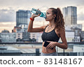 Side shot of young athletic woman drinking water from sport bottle while listening to music on city background 83148167