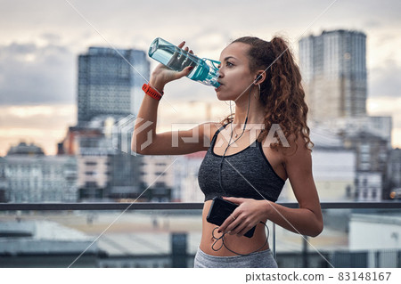 Side shot of young athletic woman drinking water from sport bottle while listening to music on city background Side shot of young athletic woman drinking water from sport bottle while listening to music on city background 83148167