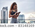 Young athletic woman in sport clothes standing on city background with phone in hands posting her training results 83148169