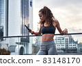 Young athletic woman in sport clothes standing on city background with phone in hands posting her training results 83148170
