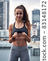 Young athletic woman in sport clothes standing on city background with phone in hands posting her training results 83148172