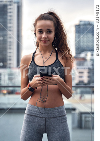 Young athletic woman in sport clothes standing on city background with phone in hands posting her training results 83148172