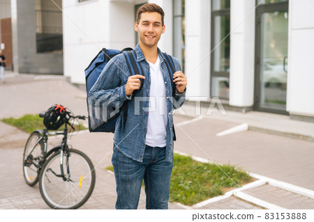 Medium shot portrait of happy handsome young delivery man with large thermo backpack standing on city street. 83153888
