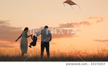 Happy family day, father mother and little kid fly a kite into the sky at sunset, play with child in nature in the evening, love spending time with family, raising and developing baby, cheerful life. Happy family day, father mother and little kid fly a kite into the sky at sunset, play with child in nature in the evening, love spending time with family, raising and developing baby, cheerful life. 83155837