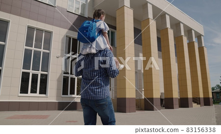 Dad carries on his shoulders little kid with backpack to school, girl with school bag rides her father around the schoolyard, happy family an elementary school, first grader with parent goes to class Dad carries on his shoulders little kid with backpack to school, girl with school bag rides her father around the schoolyard, happy family an elementary school, first grader with parent goes to class 83156338