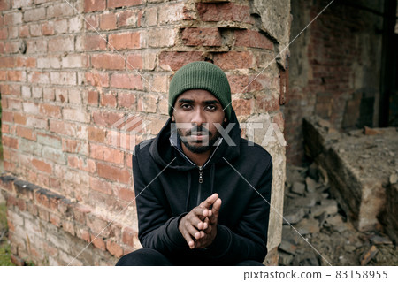 Young African man in hoodie and cap sitting by brick wall 83158955