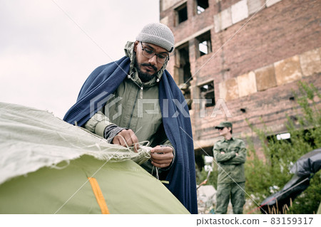 Male refugee pitching tent against half ruined building Male refugee pitching tent against half ruined building 83159317