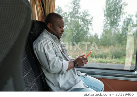 Side view of young Afrtican man with smartphone sitting by window Side view of young Afrtican man with smartphone sitting by window 83159400