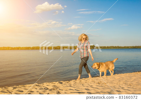 Happy young woman walking along a beach with her golden retriever at the sunset. Sun flare Happy young woman walking along a beach with her golden retriever at the sunset. Sun flare 83160372