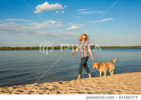 Happy young woman walking along a beach with her golden retriever at the sunset Happy young woman walking along a beach with her golden retriever at the sunset 83160373