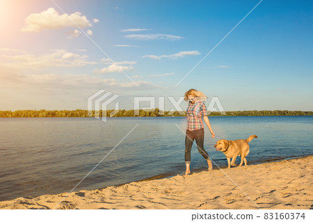 Happy young woman walking along a beach with her golden retriever at the sunset. Sun flare Happy young woman walking along a beach with her golden retriever at the sunset. Sun flare 83160374