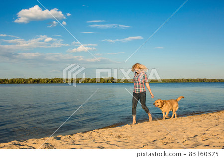 Happy young woman walking along a beach with her golden retriever at the sunset 83160375