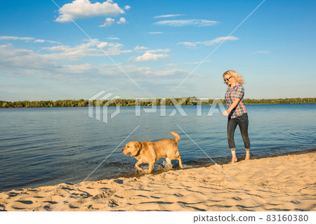 Happy young woman walking along a beach with her golden retriever at the sunset Happy young woman walking along a beach with her golden retriever at the sunset 83160380