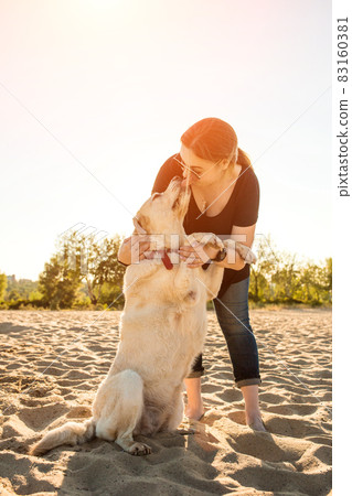 Portrait of young beautiful woman in sunglasses sitting on sand beach with golden retriever dog. Girl with dog by sea. Sun flare Portrait of young beautiful woman in sunglasses sitting on sand beach with golden retriever dog. Girl with dog by sea. Sun flare 83160381