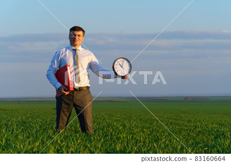 a businessman holds an office clock and red folder in a field with green grass - business and time concept a businessman holds an office clock and red folder in a field with green grass - business and time concept 83160664