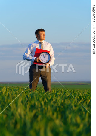 a businessman holds an office clock and red folder in a field with green grass - business and time concept a businessman holds an office clock and red folder in a field with green grass - business and time concept 83160665