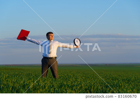 a businessman holds an office clock and red folder in a field with green grass - business and time concept a businessman holds an office clock and red folder in a field with green grass - business and time concept 83160670