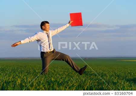 businessman walks through a green grass field and holds an office red folder with documents - business concept businessman walks through a green grass field and holds an office red folder with documents - business concept 83160704