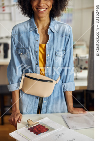 Smiling African-American clothes designer stands at table with drawings in sewing workshop Smiling African-American clothes designer stands at table with drawings in sewing workshop 83162469