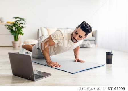 Young Arab man standing in plank pose or doing push ups near laptop, working out to online sports tutorial at home Young Arab man standing in plank pose or doing push ups near laptop, working out to online sports tutorial at home 83165909