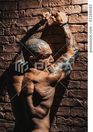 A criminal in tattoos stands against a brick wall with handcuffs on his hands. A dramatic portrait A criminal in tattoos stands against a brick wall with handcuffs on his hands. A dramatic portrait 83169083
