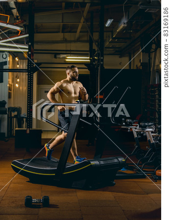 Full-length portrait of young sportive man running on a treadmill. Doing cardio. Side view Full-length portrait of young sportive man running on a treadmill. Doing cardio. Side view 83169186