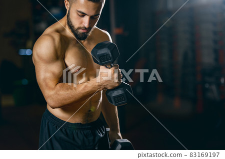 Cropped portrait of young sportive man training, pumping hands with dumbbells in gym Cropped portrait of young sportive man training, pumping hands with dumbbells in gym 83169197