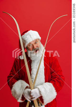 Cropped portrait of joyful senior man wearing Santa Claus costume, holding skis isolated over red background. Winter holiday season 83169346
