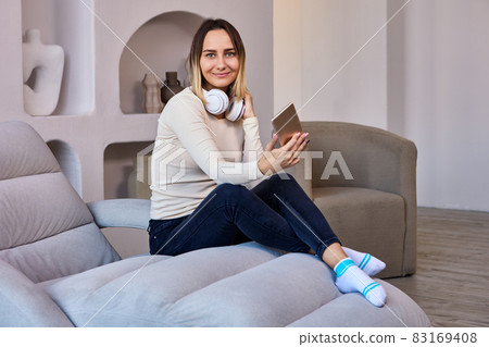 Lounge where woman on sofa and in headphones holds phone. 83169408