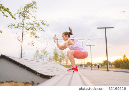 Side view of young Caucasian fit active woman jumping on bench in city park in summer morning. 83169626