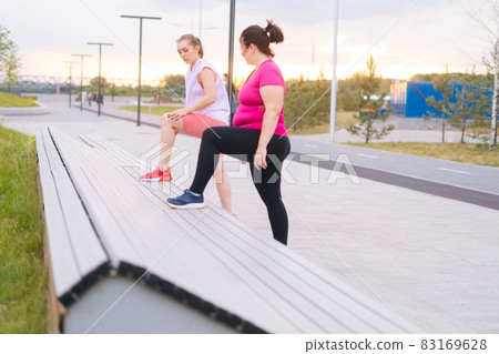 Overweight young woman doing single leg squats using bench with personal trainer outdoor in summer morning. Overweight young woman doing single leg squats using bench with personal trainer outdoor in summer morning. 83169628