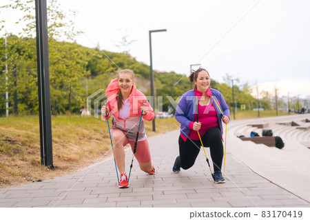 Front view of smiling fat young woman doing squats exercises using fitness tape for weight loss with personal trainer outdoor in summer day. Front view of smiling fat young woman doing squats exercises using fitness tape for weight loss with personal trainer outdoor in summer day. 83170419
