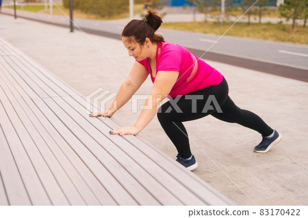 High-angle view of motivated fat young woman training doing push ups using street bench in city park at summer morning. High-angle view of motivated fat young woman training doing push ups using street bench in city park at summer morning. 83170422