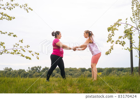 Wide shot of professional fitness female trainer giving personal training to overweight young woman outdoor in summer day. Wide shot of professional fitness female trainer giving personal training to overweight young woman outdoor in summer day. 83170424