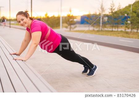Side view of smilingfat young woman training doing push ups on street bench in city park at summer morning, looking at camera. Side view of smilingfat young woman training doing push ups on street bench in city park at summer morning, looking at camera. 83170426